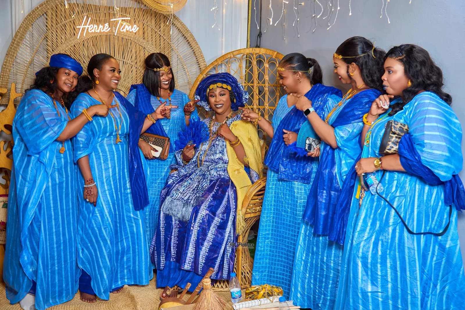 Photo de groupe, Henné Time : la mariée en bleu et wax, châle jaune, amies en bleu, décor en rotin.